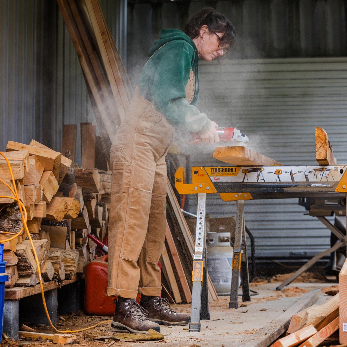 A woman dressed in overalls and brown leather boots uses a rotary sander on a rough cut wood 2x6 resting on saw horses in a covered workshop.