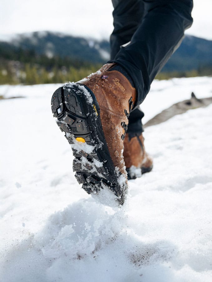 A stylishly dressed person with a backpack casually slung over one shoulder and tidily tied boots sits on a weathered wooden bench on a sunny day, the scene suggests they feel prepared for whatever their day has in store