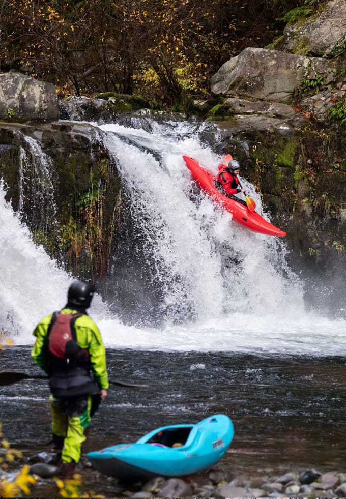 a kayaker watches from shore while another kayak goes down a waterfall