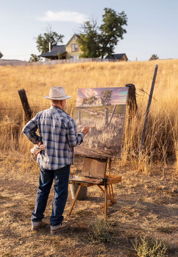a man paints while looking at a field and house