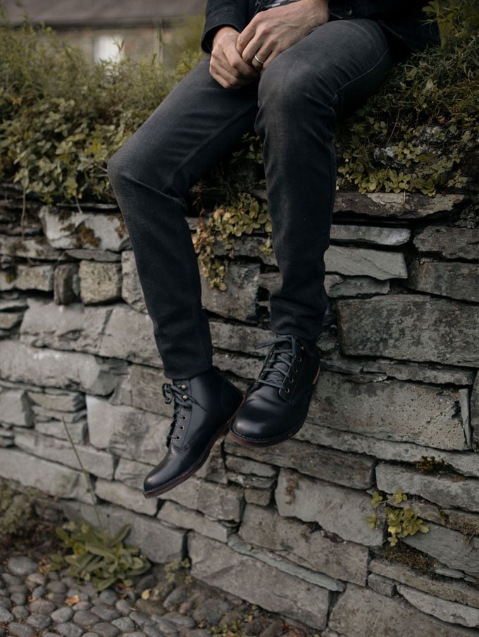 A pair of light brown leather boots with red laces rest on a pine bough with a metal mug and silverware nearby, suggesting a restful moment at camp after a hike