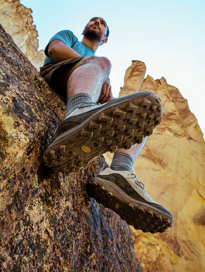 A hiker climbs rocky terrain with clouds and trees visible in the background below