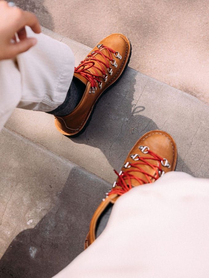 A stylishly dressed person with a backpack casually slung over one shoulder and tidily tied boots sits on a weathered wooden bench on a sunny day, the scene suggests they feel prepared for whatever their day has in store