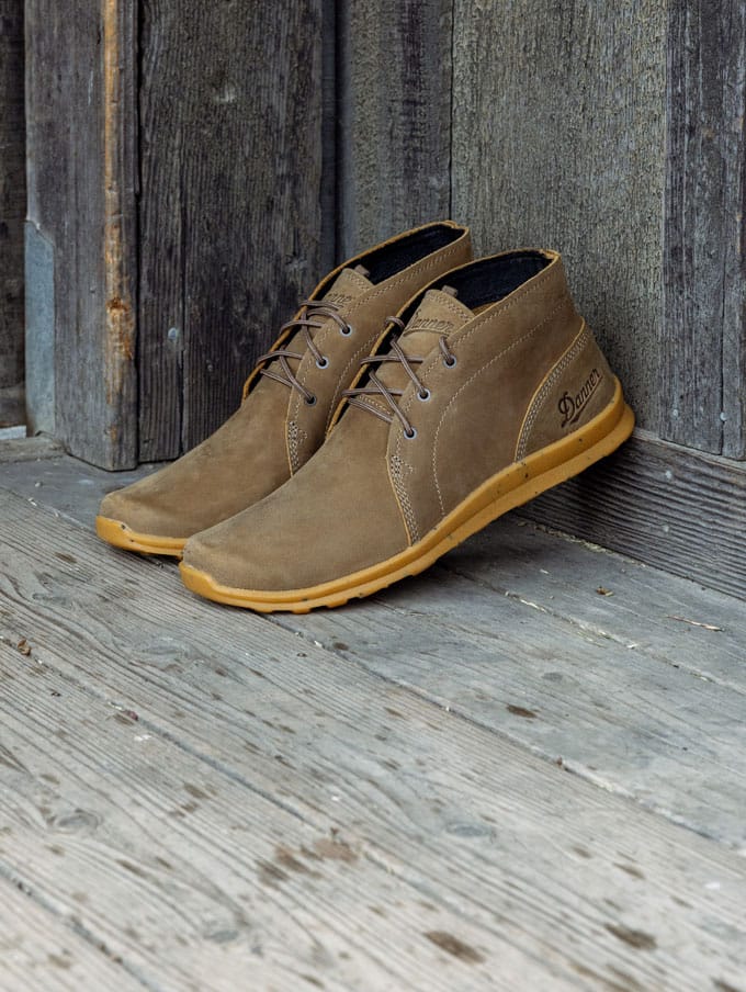 A stylishly dressed person with a backpack casually slung over one shoulder and tidily tied boots sits on a weathered wooden bench on a sunny day, the scene suggests they feel prepared for whatever their day has in store