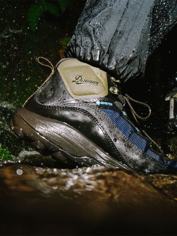 A hiker climbs rocky terrain with clouds and trees visible in the background below