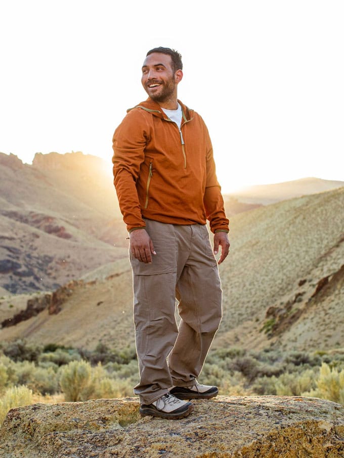 Two people walk on a well-maintained loamy trail through a lush, dark forest wearing earth-toned classically styled clothing and boots, their calm, smiling faces suggest they’re enjoying their walk and each other