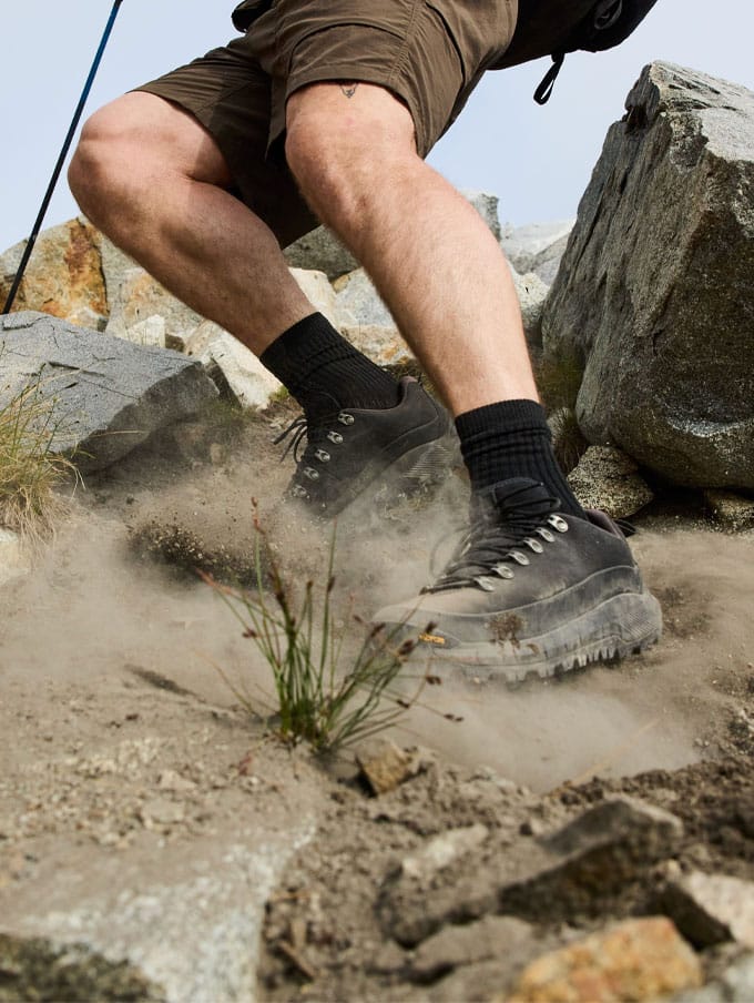 A hiker climbs rocky terrain with clouds and trees visible in the background below