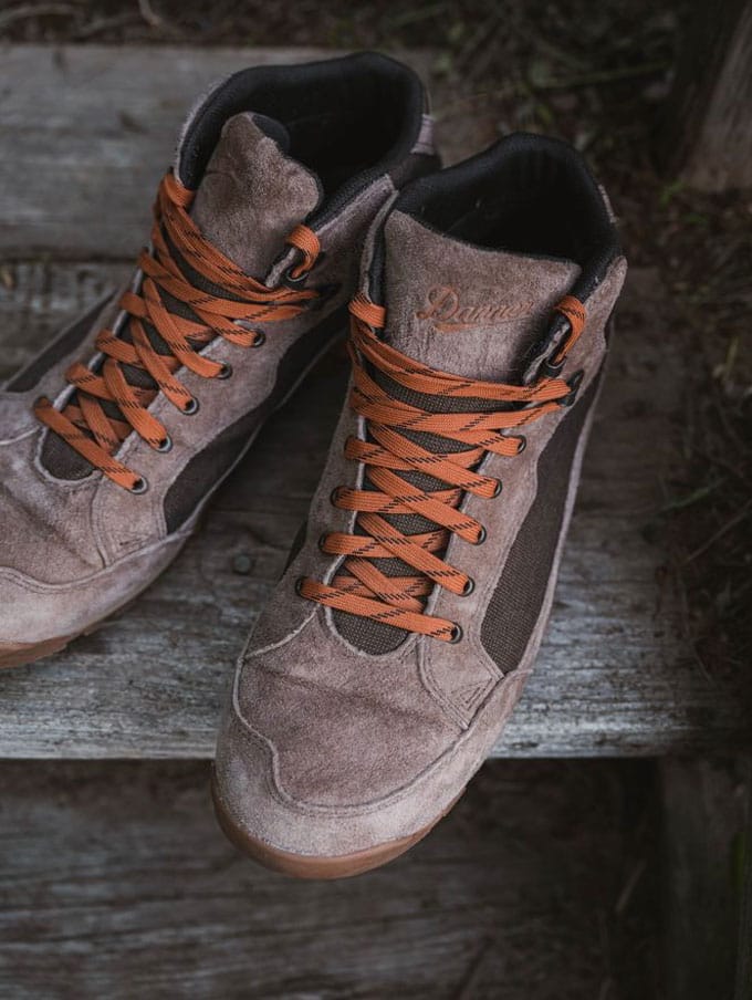 A stylishly dressed person with a backpack casually slung over one shoulder and tidily tied boots sits on a weathered wooden bench on a sunny day, the scene suggests they feel prepared for whatever their day has in store