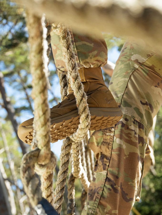 A stong-looking person suspends from a thick rope wearing camo pants, the rope is weaved around one foot showing the grippy outsole tread suggesting their boots are a key tool for their job