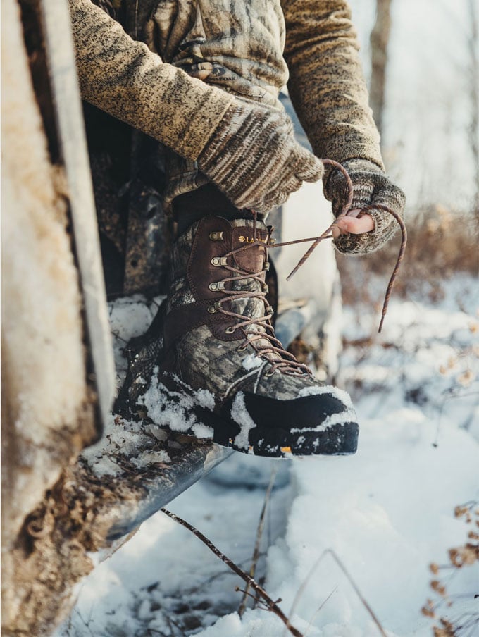 a man in a camo jacket walks through a stream while wearing brown leather hunting boots with a black rubber rand