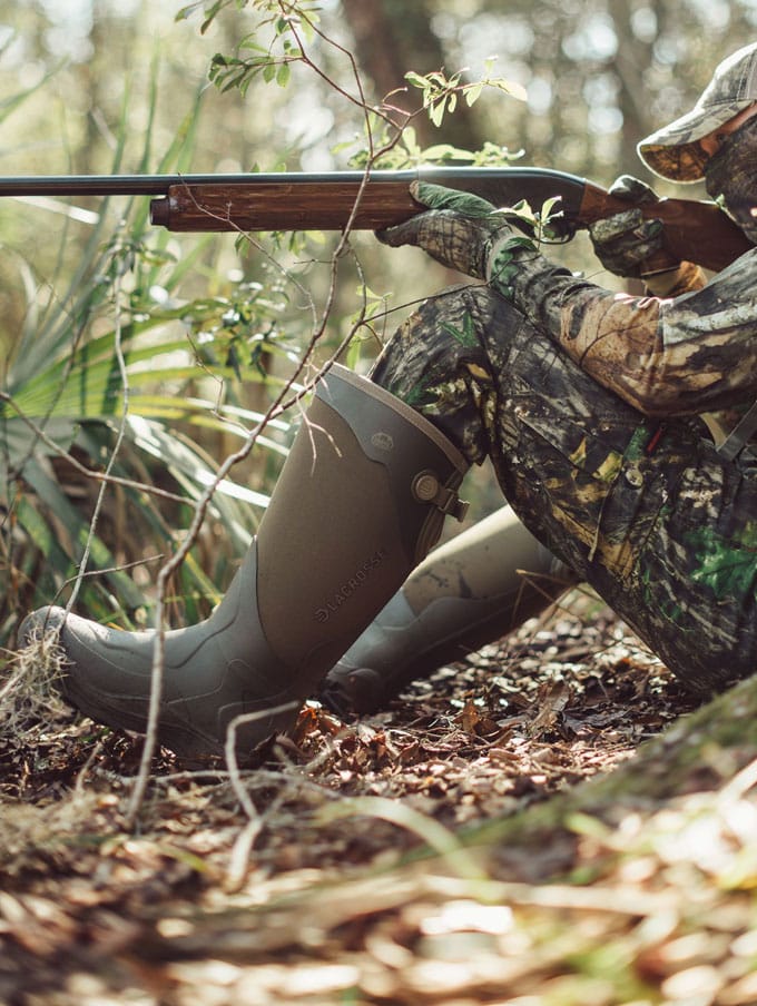a man in camo sits on the porch of a cabin while lacing up his knee high leather and nylon snake boots