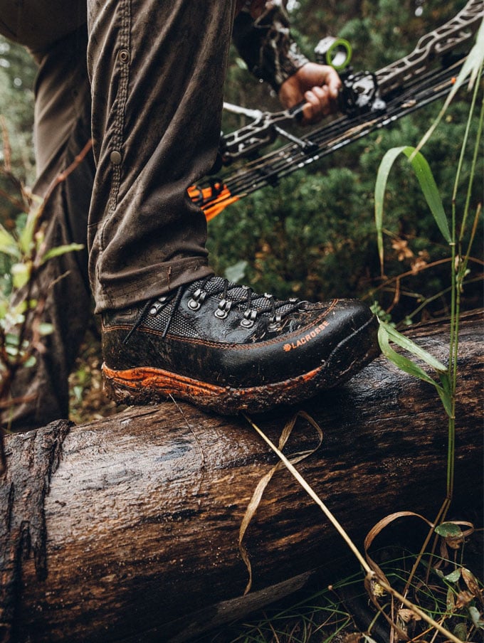 a man in a camo jacket walks through a stream while wearing brown leather hunting boots with a black rubber rand