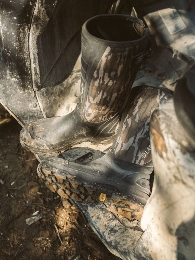 a man in camo sits on the porch of a cabin while lacing up his knee high leather and nylon snake boots