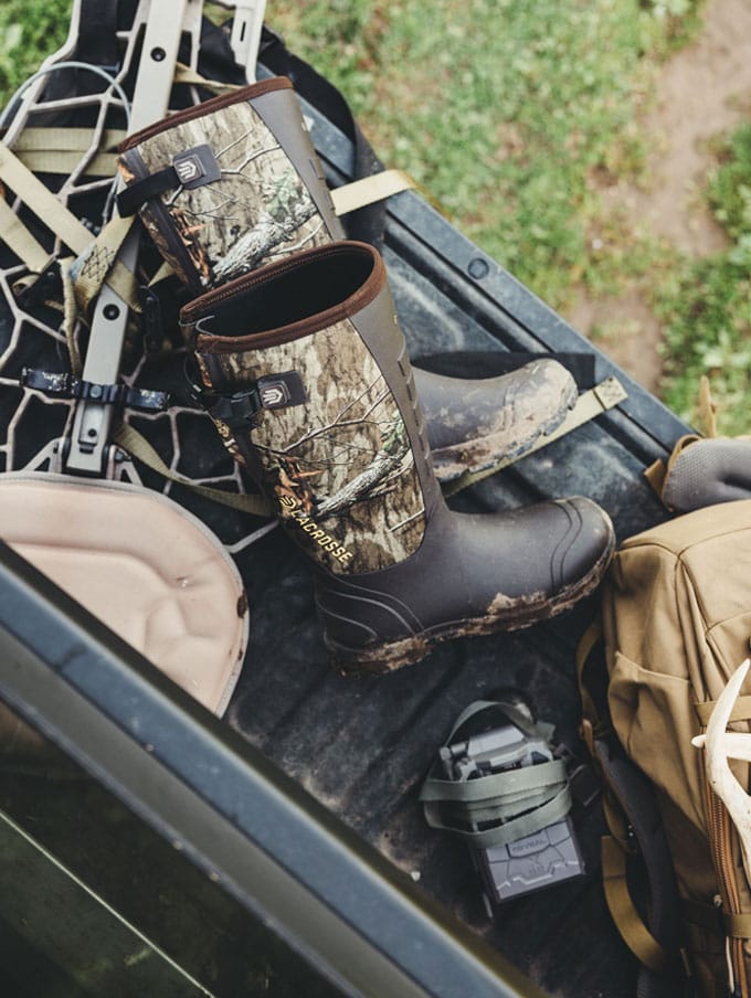 a man in camo sits on the porch of a cabin while lacing up his knee high leather and nylon snake boots