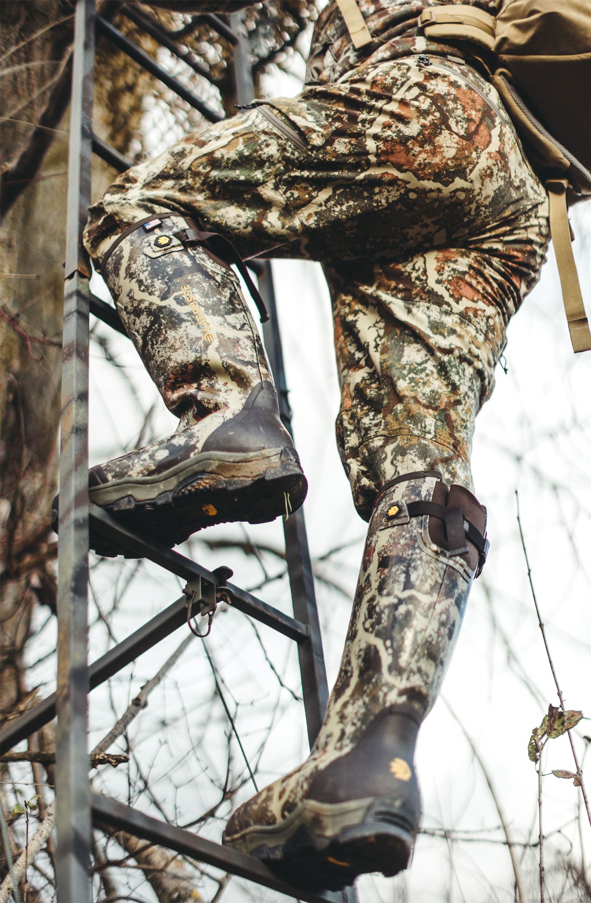 A man perched in a tree wearing gray hunting boots